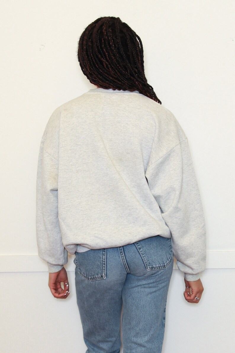 May include: A gray sweatshirt with long sleeves and a crew neck. The sweatshirt is worn by a person with long black braids. The person is wearing blue jeans.