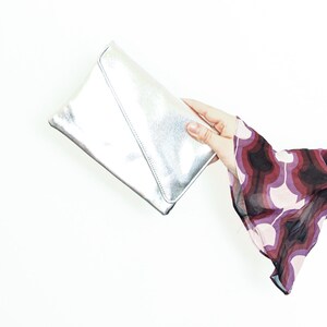 Op de afbeelding: Een zilveren metallic clutch tasje vastgehouden in een hand met een roze en paarse gepatenteerde mouw.