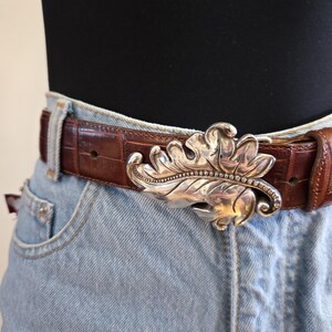 Peut inclure: Une ceinture en cuir marron avec une boucle argent&eacute;e en forme de feuille. La boucle pr&eacute;sente des d&eacute;tails complexes et une rang&eacute;e de petits ornements. La ceinture est port&eacute;e avec un jean en denim bleu clair.