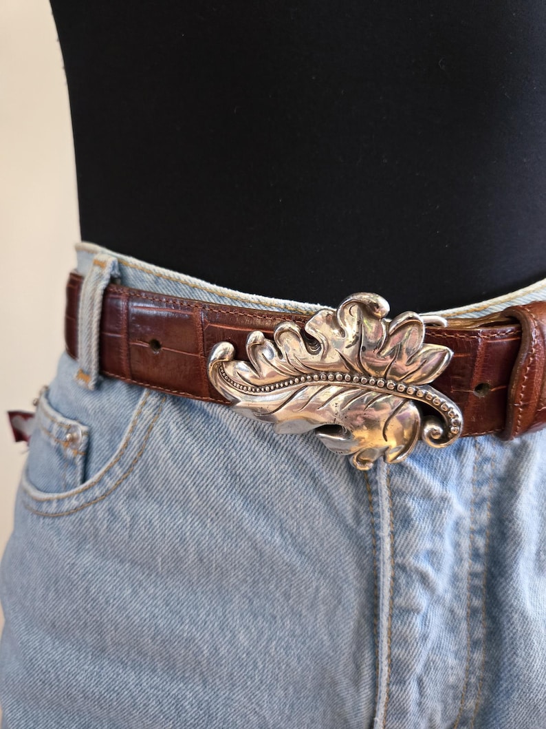 Peut inclure: Une ceinture en cuir marron avec une boucle argent&eacute;e en forme de feuille. La boucle pr&eacute;sente des d&eacute;tails complexes et une rang&eacute;e de petits ornements. La ceinture est port&eacute;e avec un jean en denim bleu clair.