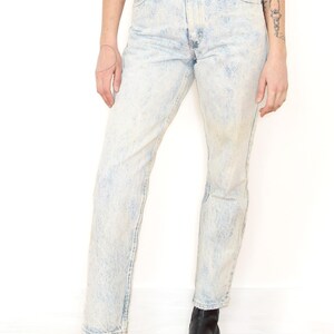 May include: High-waisted denim jeans featuring a light blue acid wash finish, a classic five-pocket design, a metal button closure at the waist, and a straight-leg cut that extends to the ankles, paired with black leather ankle boots.