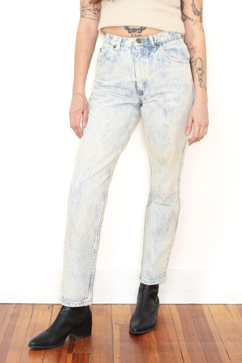 May include: High-waisted denim jeans featuring a light blue acid wash finish, a classic five-pocket design, a metal button closure at the waist, and a straight-leg cut that extends to the ankles, paired with black leather ankle boots.