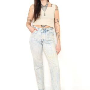 May include: A beige sleeveless crop top with a square neckline paired with high-waisted acid wash blue jeans featuring a light mottled pattern, a button fly, and straight legs, worn with black leather ankle boots and a silver chainmail necklace.