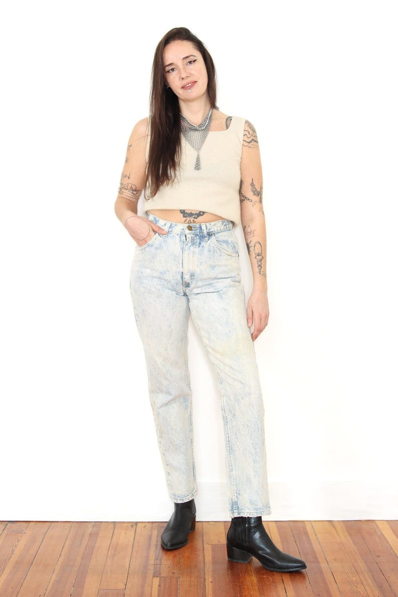 May include: A beige sleeveless crop top with a square neckline paired with high-waisted acid wash blue jeans featuring a light mottled pattern, a button fly, and straight legs, worn with black leather ankle boots and a silver chainmail necklace.