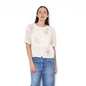 May include: A cream-colored short-sleeve knit cardigan featuring a button-down front, a delicate openwork diamond pattern, and embroidered floral motifs in shades of pink, blue, and yellow, paired with blue denim jeans.
