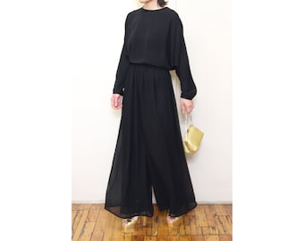 May include: A black long-sleeved jumpsuit with a gathered waist and wide legs. The jumpsuit is made of a lightweight fabric and has a relaxed fit. The model is wearing gold sandals and carrying a gold clutch.