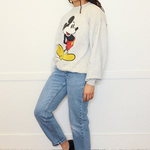 May include: A gray sweatshirt with a Mickey Mouse graphic. The sweatshirt has long sleeves and a crew neck. The person wearing the sweatshirt is also wearing blue jeans and black shoes.