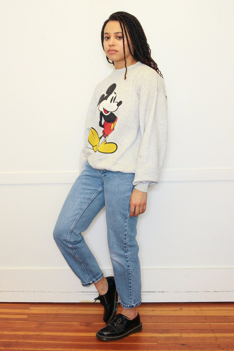 May include: A gray sweatshirt with a Mickey Mouse graphic. The sweatshirt has long sleeves and a crew neck. The person wearing the sweatshirt is also wearing blue jeans and black shoes.