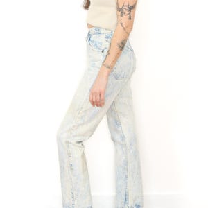 May include: High-waisted acid wash denim jeans in a light blue and white mottled pattern. The jeans feature a classic five-pocket design with a straight leg cut, a button fly closure, and visible stitching along the seams, ending at the ankle.
