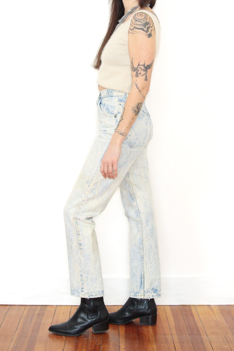 May include: High-waisted acid wash denim jeans in a light blue and white mottled pattern. The jeans feature a classic five-pocket design with a straight leg cut, a button fly closure, and visible stitching along the seams, ending at the ankle.