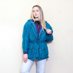 Könnte beinhalten: Eine türkisblaue Kapuzenjacke mit Kordelzug in der Taille und zwei Fronttaschen. Die Jacke hat einen Reißverschluss und eine Knopfleiste über dem Reißverschluss.