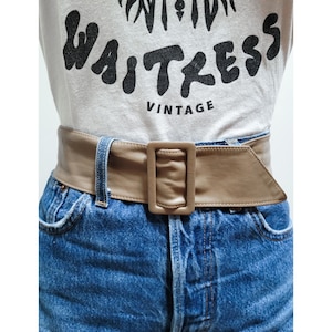 Puede incluir: Un cinturón ancho de cuero marrón con una hebilla cuadrada grande. El cinturón se lleva sobre unos vaqueros azules.