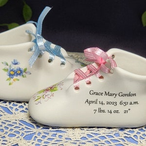 May include: Two white ceramic baby shoes with blue and pink ribbons. The shoes are decorated with floral designs. The larger shoe has the text "Grace Mary Gordon April 14, 2023 6:51 a.m. 7 lbs. 14 oz. 21"" inscribed on the side.