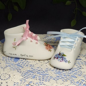 May include: Two porcelain baby shoes with pink and blue ribbons. The shoes are decorated with floral designs and the pink shoe has the inscription "7:55 a.m. ... April 14, 2021".
