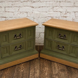 May include: A pair of matching nightstands featuring two drawers each, finished in a muted olive green paint with natural light wood tops and bases. Each drawer is accented with decorative recessed panels and ornate, curved metal pull handles.