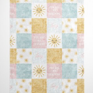 May include: A pastel pink, blue, and yellow patchwork baby blanket with a sun and the name "Lucy" repeated throughout. The blanket has the text "You are my sunshine" on it.