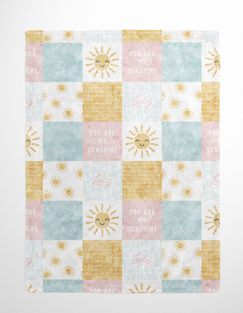 May include: A pastel pink, blue, and yellow patchwork baby blanket with a sun and the name "Lucy" repeated throughout. The blanket has the text "You are my sunshine" on it.