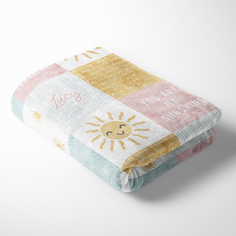 May include: A soft, plush blanket with a patchwork design featuring a smiling sun, stripes, polka dots, and the text "You are my sunshine". The blanket is pink, yellow, blue, and white.