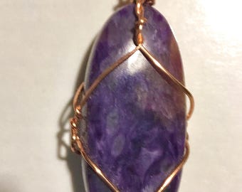 May include: A handcrafted pendant featuring a polished, oval-shaped purple stone. The stone is wrapped in copper wire, creating a cage-like design. The wire is intricately woven at the top to form a loop for hanging. The pendant has a natural, organic aesthetic.