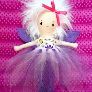 May include: A handmade fairy doll with white fluffy hair, a pink ribbon, and purple wings. The doll wears a white dress with floral details and a layered purple and white tulle skirt. The background is a pink fabric with a heart pattern.