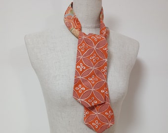 May include: A necktie featuring a vibrant orange background with a repeating white and muted pink geometric pattern of interlocking circles and small four-petaled floral shapes. The tie is tied in a simple knot around a neutral-colored mannequin neck.