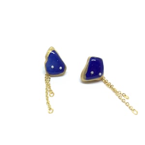 Könnte beinhalten: Ein Paar einzigartige Ohrringe mit unregelmäßigen, tiefblauen Formen, goldenen Rändern und kleinen weißen Akzenten. An jedem Ohrring hängen zarte Goldketten, die ein stilvolles und künstlerisches Accessoire bilden.