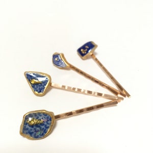 May include: Four decorative hair pins with blue and gold detailing. Each pin showcases a unique design, including fish and floral patterns. The pins have a gold-coloured metal base and are set against a white background.