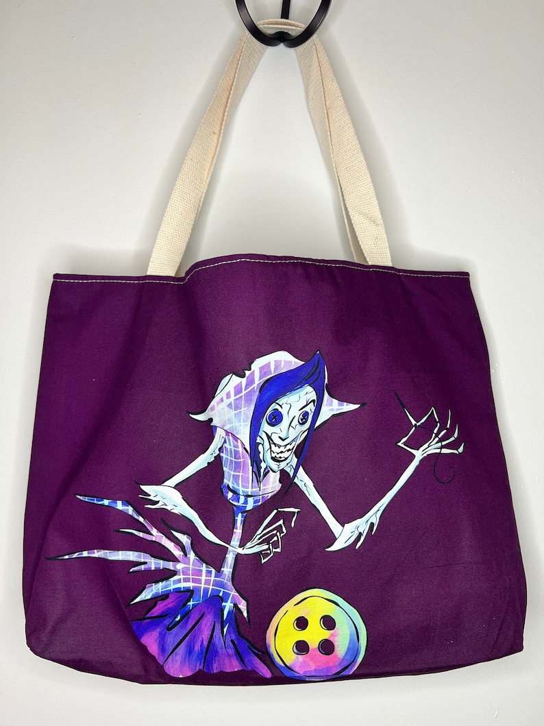 Pu&ograve; includere: Borsa tote viola con una tracolla in tela bianca. La borsa presenta un'illustrazione dipinta a mano di un personaggio dei cartoni animati con un grande bottone e un ago.