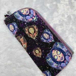 May include: A rectangular, zippered pencil case featuring a repeating pattern of a cartoon character with a skull-like face, surrounded by floral and decorative elements. The background is black with a purple web-like design.