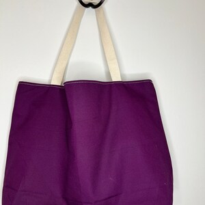 Pu&ograve; includere: Una borsa tote viola con manici color crema appesa a un gancio.