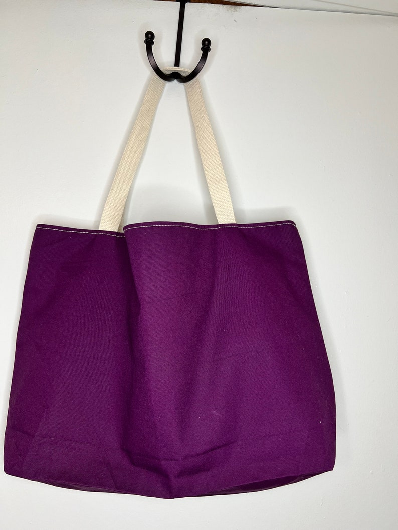 Pu&ograve; includere: Una borsa tote viola con manici color crema appesa a un gancio.