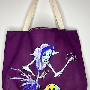 Pu&ograve; includere: Borsa tote viola con un'illustrazione bianca e blu di una creatura con braccia lunghe e un grande bottone. La creatura ha una testa grande con un ampio sorriso e un naso a forma di bottone.