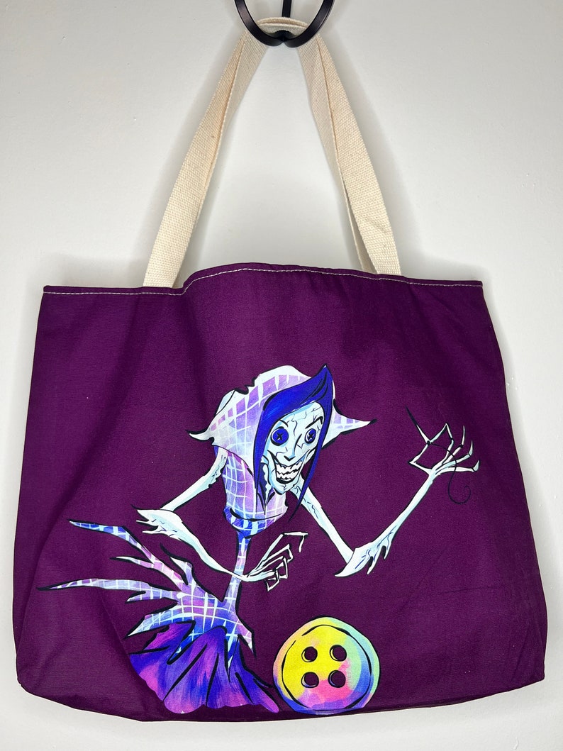 Pu&ograve; includere: Borsa tote viola con un'illustrazione bianca e blu di una creatura con braccia lunghe e un grande bottone. La creatura ha una testa grande con un ampio sorriso e un naso a forma di bottone.
