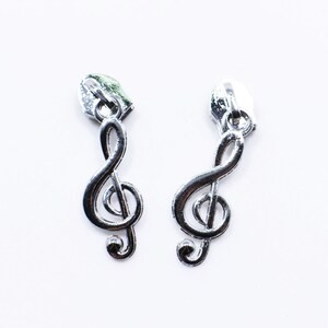 以下が含まれることがあります： 2つの銀色のジッパープルで、それぞれにト音記号のデザインが施されています。ジッパープルは金属製のトップから吊り下げられており、音楽記号は曲線で詳細に描かれています。背景は無地の白です。