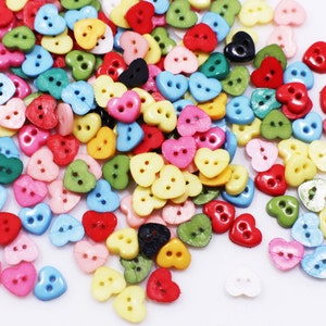 May include: A large assortment of colourful heart-shaped buttons. The buttons are made of plastic and have two holes. The buttons are in a variety of colours, including red, pink, blue, green, yellow, and white.