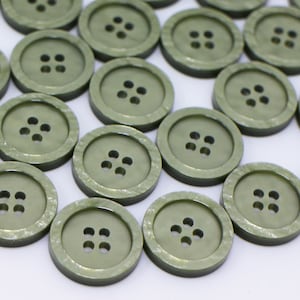 May include: A selection of olive green, circular buttons, each with four holes. The buttons are arranged in a scattered pattern, highlighting their consistent design and colour. These buttons are suitable for various sewing projects.