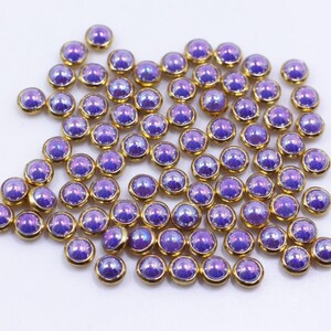 Peut inclure: Un grand groupe de petits strass ronds, violets irisés, avec des montures dorées. Les strass sont éparpillés sur une surface blanche.