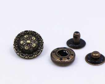 May include: A set of three antique gold-toned metal snap fasteners. The largest snap has a decorative, ornate design. The other two snaps are smaller and have a more simple design.