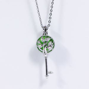 May include: Silver-toned key pendant necklace with a green, tree-shaped design inside a round pendant. The key pendant hangs from a delicate chain. The necklace is displayed against a white background.