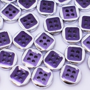 Peut inclure: Un grand groupe de boutons violets avec une bordure argentée et quatre trous chacun. Les boutons sont éparpillés sur une surface blanche.
