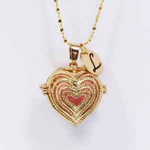 Könnte beinhalten: Goldene Halskette mit herzförmigem Anhänger, einer zarten Kette und einem kleinen Anhänger mit dem Buchstaben 'L'. Der Anhänger hat ein rosafarbenes Emaille-Innere und komplizierte Details.