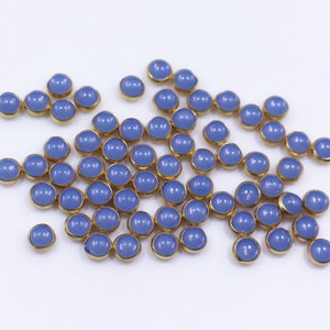May include: A large group of small, round, blue glass beads with gold trim. The beads are scattered on a white surface.