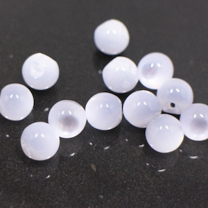 May include: A group of white glass beads with a slightly iridescent finish. The beads are scattered on a black surface.