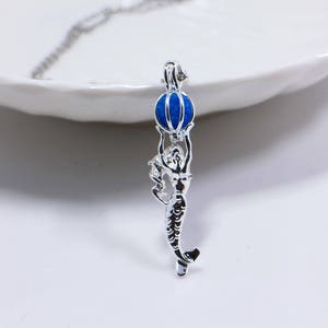 Peut inclure: Collier en argent avec une sirène tenant une boule bleue et blanche à rayures au-dessus de sa tête.