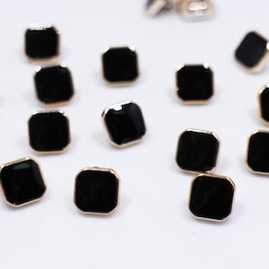 May include: A collection of black square buttons with gold trim. The buttons are scattered on a white surface.