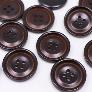 May include: A collection of brown plastic buttons with four holes each. The buttons are arranged in a random pattern on a white surface.