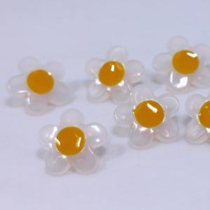 May include: A group of small, white plastic daisy buttons with yellow centres. The buttons are scattered on a white surface.