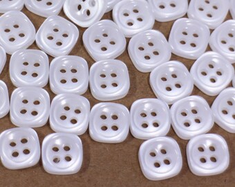 May include: A large quantity of white plastic buttons with four holes each. The buttons are square shaped with rounded corners.