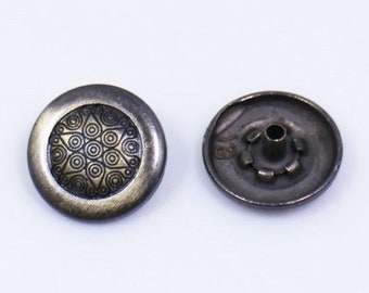 May include: A pair of round, metal buttons with a decorative design. The button on the left has a gold-colored, circular pattern. The button on the right is plain and has a small hole in the center.