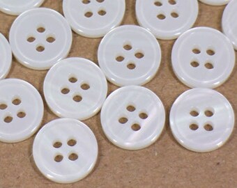 May include: A collection of white, round buttons with four holes each. The buttons are scattered across a brown surface. These sewing notions are ideal for various craft and apparel projects.